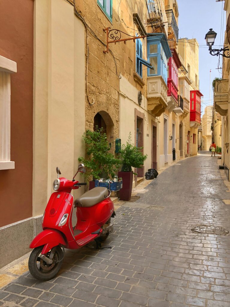 Quaint Maltese street with colorful balconies and a parked red Vespa, showcasing historic architecture.