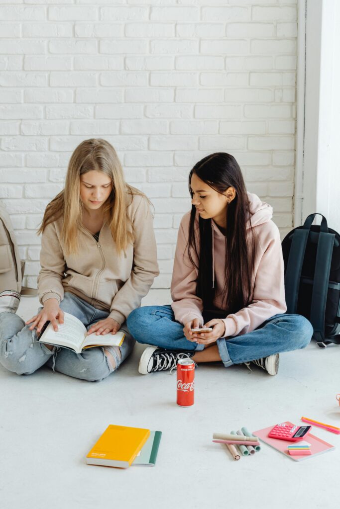 Two teenagers studying indoors with books and stationery on a white brick background.