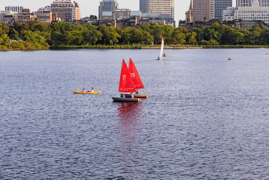 Red sailboats and a kayak on Charles River with Boston skyline in the background, capturing a sunny day.