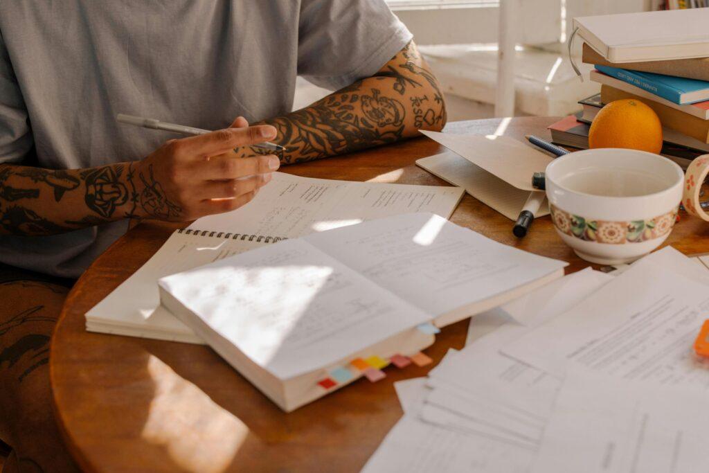 A tattooed adult studying with notes and textbooks at a sunlit wooden desk.