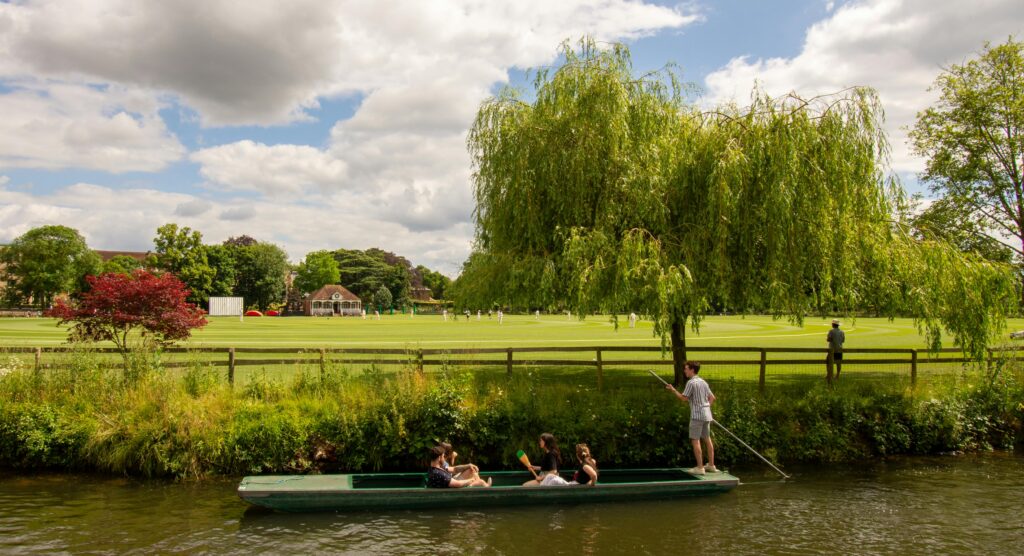 Enjoy a relaxing punting experience on the river in Oxford with lush greenery and a cricket field backdrop.