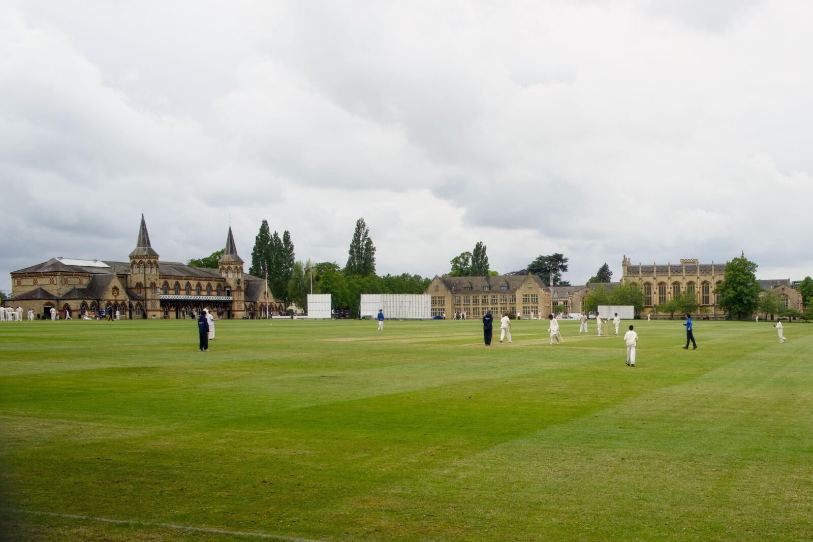 Outdoor cricket match on a lush green field with a historic building backdrop, overcast sky, many players visible.